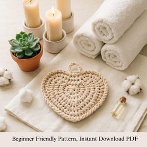 May include: A beige, heart-shaped, crocheted item with a loop for hanging. The image includes rolled white towels, lit candles in white holders, a small potted succulent, cotton buds, and a small glass bottle. Text at the bottom reads: Beginner Friendly Pattern, Instant Download PDF.