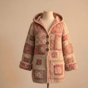 May include: A hooded, long-sleeved crochet jacket in a patchwork design. The jacket features a mix of light pink and cream-colored squares and floral patterns. It has three brown buttons down the front and rolled-up cuffs.