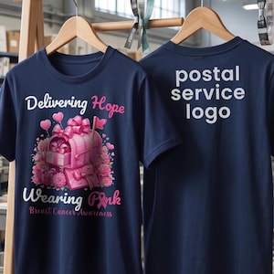 May include: A navy blue t-shirt featuring a graphic of a pink mailbox surrounded by flowers and gifts, with the text Delivering Hope Wearing Pink Breast Cancer Awareness. The back of the shirt displays the text postal service logo in white lettering.