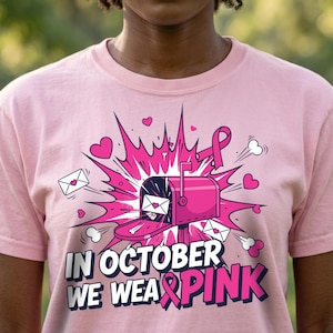 May include: A light pink t-shirt featuring a graphic of a bright pink mailbox surrounded by pink hearts, envelopes, and a pink ribbon. The text "IN OCTOBER WE WEAR PINK" is printed in bold white and pink letters below the mailbox illustration.