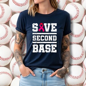 May include: Navy blue t-shirt with the text "SAVE SECOND BASE" in white block letters. A pink ribbon is incorporated into the word "SAVE". The shirt is a baseball-themed design.