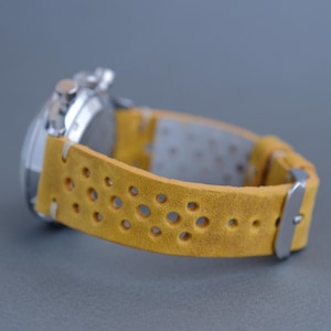 Könnte beinhalten: Nahaufnahme einer Armbanduhr mit silbernem Gehäuse und gelbem Lederarmband. Das Armband hat ein perforiertes Design und eine silberne Schnalle. Die Uhr ist vor einem grauen Hintergrund platziert.