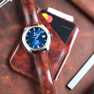 Könnte beinhalten: Eine silberne Rado-Uhr mit blauem Zifferblatt und braunem Lederarmband liegt auf einem braunen Lederportemonnaie. Das Zifferblatt zeigt das Rado-Logo und die Worte "Golden Horse". Eine schwarze Kreditkarte ist im Portemonnaie sichtbar.