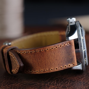Op de afbeelding: Close-up van een bruin leren horlogebandje met zichtbare stiksels. De band is bevestigd aan een zilveren horloge met een zwarte wijzerplaat en een draaibare bezel. Het leer heeft een verweerde uitstraling.