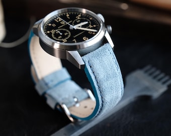 Peut inclure: Une montre chronographe en acier inoxydable avec un cadran noir doté de chiffres et de sous-cadrans blancs. La montre est équipée d'un bracelet en cuir suédé bleu clair avec des coutures visibles et une boucle en métal argenté.