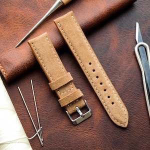 Puede incluir: Dos correas de reloj de ante marrón claro con costuras blancas y hebillas plateadas. Las correas se muestran sobre una superficie de cuero marrón, junto con herramientas de marroquinería, como agujas y un cuchillo.