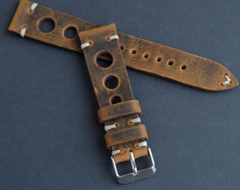 May include: Distressed brown leather watch strap with white stitching and large circular cutouts. The strap features a silver-colored buckle and a classic design, perfect for a vintage or rugged aesthetic. The watch band is approximately 9 inches long.