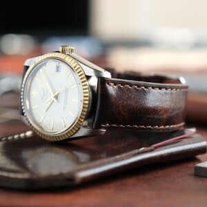 May include: A wristwatch with a silver case and a gold bezel. The watch has a white face with gold hands and markers. It features a brown leather strap. The watch is resting on a brown leather surface.