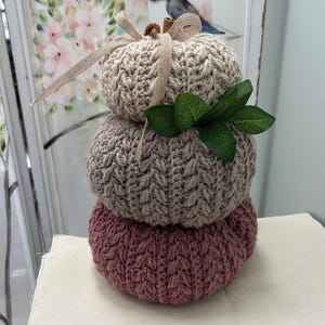 May include: A decorative stack of three knitted pumpkins in beige, grey, and mauve. The top pumpkin has a beige ribbon and a small cinnamon stick. Green leaves are tucked between the top two pumpkins. The pumpkins are on a white surface.