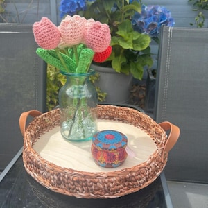 May include: A round woven tray with a wooden base and leather handles displays a glass vase of crocheted tulips. A small, decorated tin sits on the tray. Blue hydrangeas are in the background.