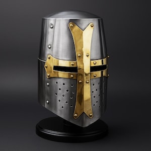May include: A silver-colored medieval crusader helmet with a gold-colored cross design. The helmet has a rounded top and is mounted on a black base. The helmet features small rivets and ventilation holes.