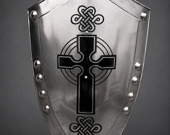 May include: A silver-colored shield with a pointed bottom and rounded top. The shield features a black Celtic cross design in the center, with decorative knotwork above and below. Rivets line the perimeter.