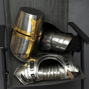 May include: A set of medieval armor, including a helmet with gold accents, shoulder and arm protection, and a gauntlet. The armor is made of polished metal and rests on a gray chair.