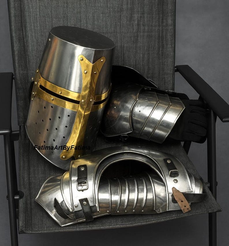 May include: A set of medieval armor, including a helmet with gold accents, shoulder and arm protection, and a gauntlet. The armor is made of polished metal and rests on a gray chair.