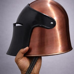 May include: A copper-colored metal helmet with a black visor. The helmet has a smooth, polished finish and a leather strap. The visor has a small opening for the eyes. The helmet is held up by a hand.