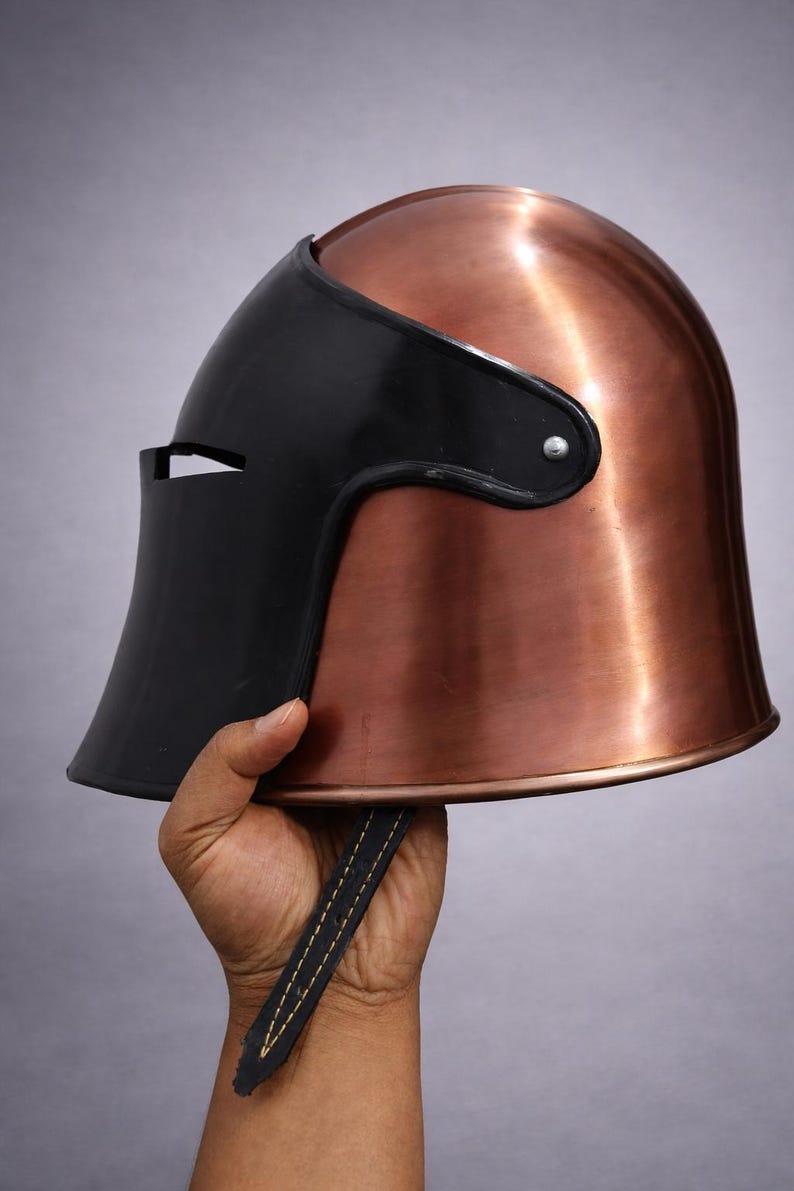 May include: A copper-colored metal helmet with a black visor. The helmet has a smooth, polished finish and a leather strap. The visor has a small opening for the eyes. The helmet is held up by a hand.