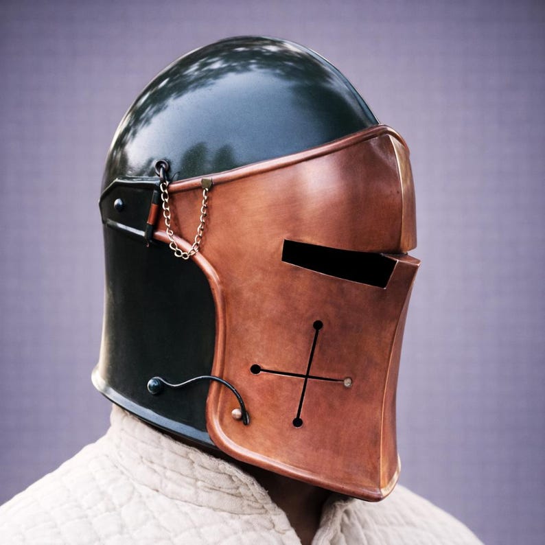 Puede incluir: Un casco de estilo medieval con un dise&ntilde;o de dos tonos. La parte superior es de color verde oscuro, mientras que la placa frontal es de color cobre con un dise&ntilde;o de cruz. Una peque&ntilde;a cadena y una pieza de metal curvada est&aacute;n unidas al lateral. El casco se lleva sobre una prenda blanca.