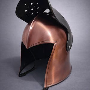 May include: A copper-colored metal helmet with a black leather visor. The helmet has a pointed front and a curved top, with a series of small holes in the visor. The helmet is likely for historical reenactment or display.