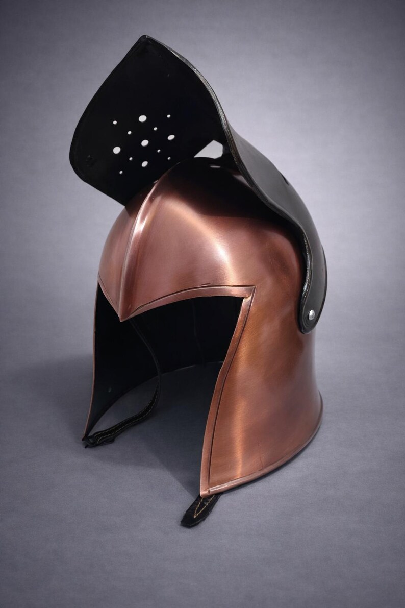 May include: A copper-colored metal helmet with a black leather visor. The helmet has a pointed front and a curved top, with a series of small holes in the visor. The helmet is likely for historical reenactment or display.