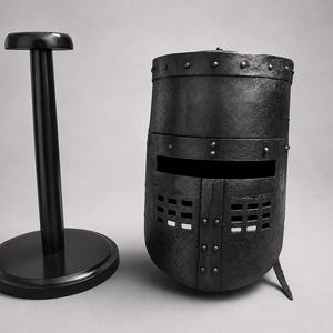 Op de afbeelding: Een zwarte middeleeuwse helm en standaard. De helm is van metaal en heeft een afgeronde vorm met een vizier en ventilatiegaten. De standaard is ook zwart, met een ronde basis en een verticale paal.