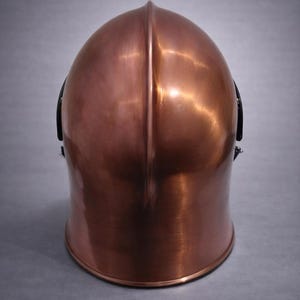May include: A copper-colored helmet with a rounded top and a vertical ridge down the center. The helmet has a polished finish, reflecting light, and black accents on the sides. The helmet is likely for protection.