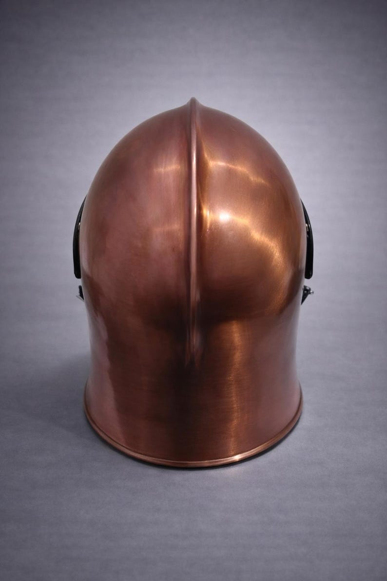 May include: A copper-colored helmet with a rounded top and a vertical ridge down the center. The helmet has a polished finish, reflecting light, and black accents on the sides. The helmet is likely for protection.