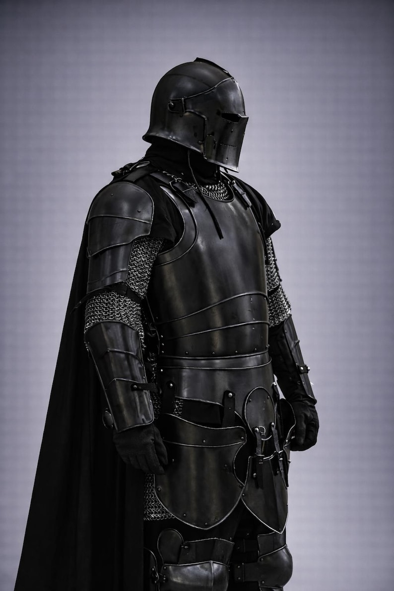 May include: A full suit of black medieval armor, including a helmet, chest plate, and leg protection. The armor features chainmail accents and a black cape. The image is a studio shot with a neutral background.