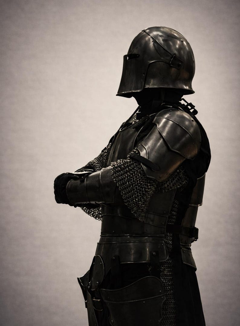May include: A full suit of medieval armor, crafted from dark metal. The armor includes a helmet, shoulder plates, chest and back protection, and leg guards. Chainmail is visible on the arms. The figure is posed with arms crossed.
