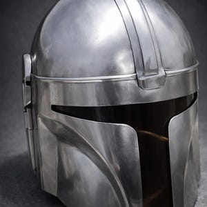 Może przedstawiać: Srebrny hełm o charakterystycznym designie, z zaokrągloną g&oacute;rą, wizjerem i wyraźnym grzebieniem brwi. Hełm ma gładkie, metaliczne wykończenie i wydaje się być wykonany z metalu. Og&oacute;lna estetyka sugeruje motyw futurystyczny lub science fiction.