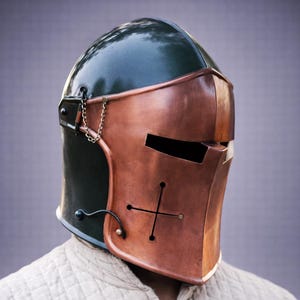 Puede incluir: Un casco de estilo medieval con una placa frontal de color cobre y una parte superior verde oscuro. El casco presenta un dise&ntilde;o de cruz y un detalle de cadena. El dise&ntilde;o recuerda a las armaduras hist&oacute;ricas.