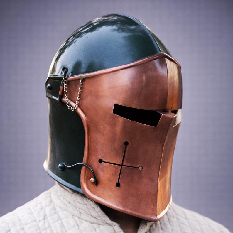 Puede incluir: Un casco de estilo medieval con una placa frontal de color cobre y una parte superior verde oscuro. El casco presenta un dise&ntilde;o de cruz y un detalle de cadena. El dise&ntilde;o recuerda a las armaduras hist&oacute;ricas.