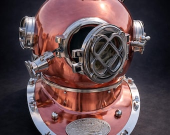 Op de afbeelding: Een vintage koperen duikhelm met zilveren accenten. De helm heeft een ronde bovenkant met een scharnierend voorvenster en diverse zilveren armaturen. Een decoratief plaatje is zichtbaar aan de voorkant. Een klassiek stuk nautische decoratie.