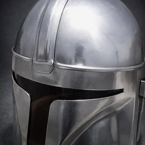Może przedstawiać: Srebrny hełm Mandalorian z gładkim, metalicznym wykończeniem. Hełm posiada charakterystyczny wizjer i zakrzywioną konstrukcję z wyraźnym grzbietem biegnącym wzdłuż g&oacute;rnej części. Og&oacute;lna estetyka sugeruje futurystyczny lub science-fiction motyw.