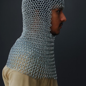 May include: A silver chainmail coif, a type of medieval head and neck protection, is shown in profile. The coif is made of small, interlocking metal rings, creating a flexible yet protective covering. The background is a solid, dark gray.
