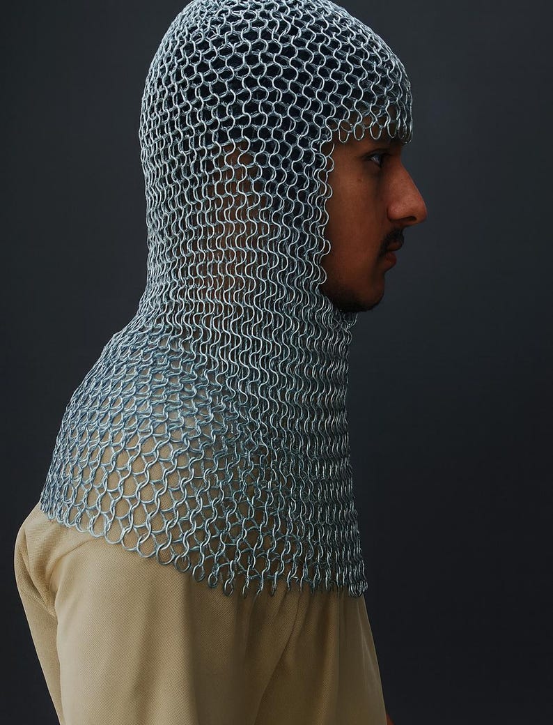 May include: A silver chainmail coif, a type of medieval head and neck protection, is shown in profile. The coif is made of small, interlocking metal rings, creating a flexible yet protective covering. The background is a solid, dark gray.