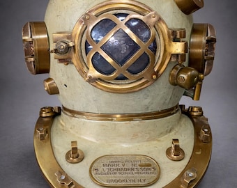 Pode incluir: Capacete de mergulho vintage, creme e latão. Apresenta uma janela frontal redonda com um design de trama cruzada e acessórios de latão. Uma placa diz "MARK V N. A. SCHRADER'S SON BROOKLYN N.Y.". Um item de colecionador.