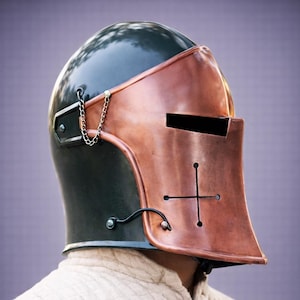 Puede incluir: Un casco de estilo medieval con una corona verde oscuro y una placa frontal de color cobre. La placa frontal presenta un dise&ntilde;o de cruz y una ranura horizontal para los ojos. Una peque&ntilde;a cadena est&aacute; unida al lateral del casco.