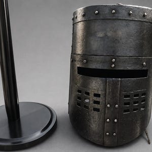 Può includere: Un elmo da cavaliere in metallo usurato, con visiera rettangolare e rivetti. L'elmo è esposto accanto a un supporto nero, suggerendo un oggetto da esposizione o da collezione. Il design evoca la storia medievale.