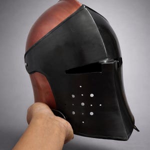 May include: A medieval-style helmet with a two-tone design. The top is a reddish-brown color, while the face guard is black with small circular ventilation holes. The helmet is held by a hand.