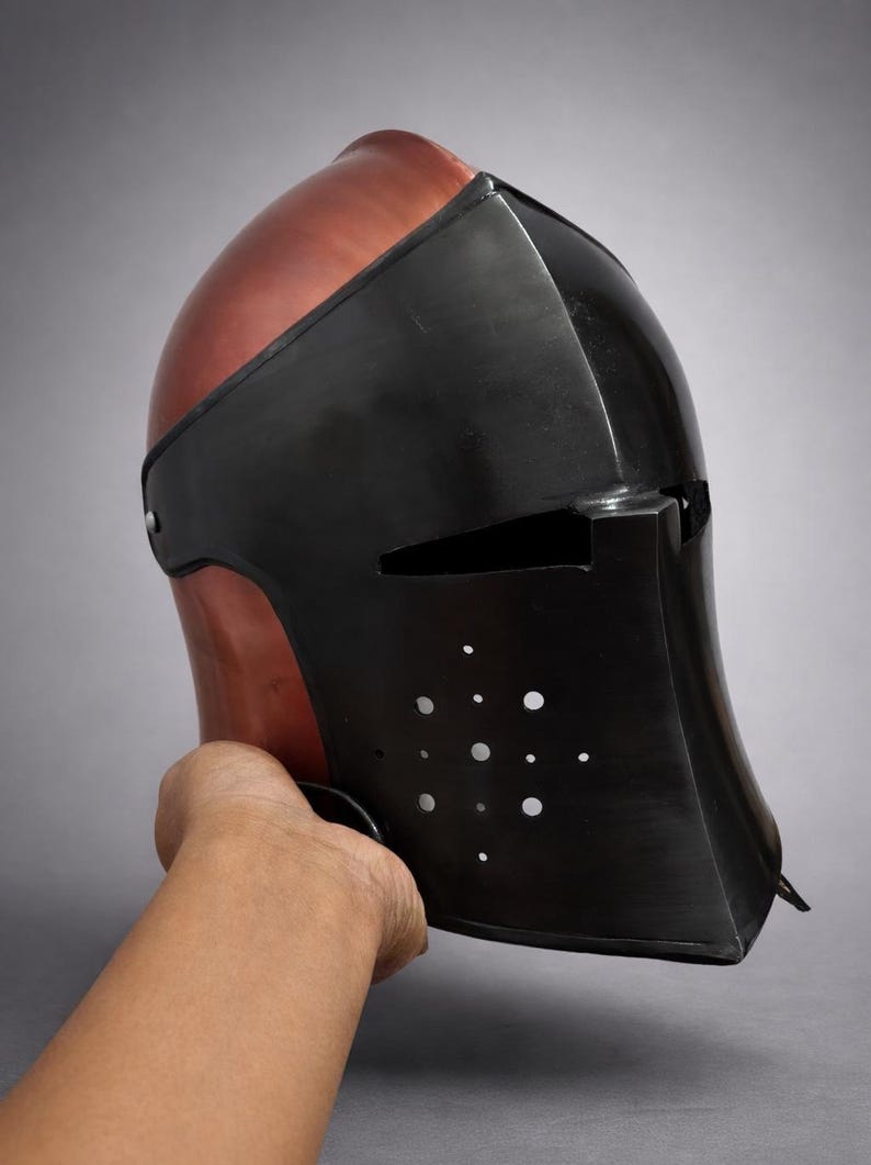 May include: A medieval-style helmet with a two-tone design. The top is a reddish-brown color, while the face guard is black with small circular ventilation holes. The helmet is held by a hand.
