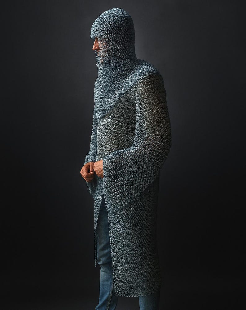 May include: A full-length, chainmail garment, including a hood, worn over blue jeans. The metallic armor is a silvery-gray color and covers the entire upper body and head. The person is standing in profile against a dark background.