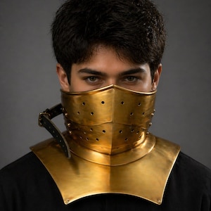 Può includere: Un gorgiera e una maschera facciale in metallo color oro. La maschera ha piccoli fori di ventilazione circolari ed è attaccata a un colletto. Il colletto è fissato con un cinturino in pelle nera. La persona indossa una camicia nera.