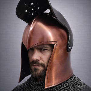 May include: A copper-colored helmet with a black visor and a chainmail collar. The helmet has a pointed top and side pieces. The visor has several small holes. The helmet is worn by a person with a beard.