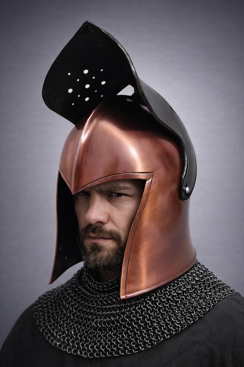 May include: A copper-colored helmet with a black visor and a chainmail collar. The helmet has a pointed top and side pieces. The visor has several small holes. The helmet is worn by a person with a beard.