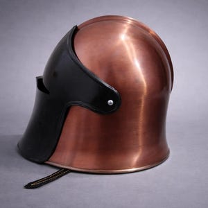 May include: A copper-colored helmet with a black visor and trim. The helmet has a smooth, polished surface and a strap at the bottom. The helmet is likely for historical reenactment or display.