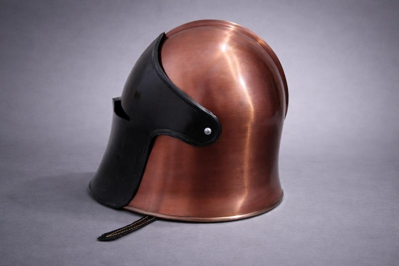 May include: A copper-colored helmet with a black visor and trim. The helmet has a smooth, polished surface and a strap at the bottom. The helmet is likely for historical reenactment or display.