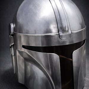Może przedstawiać: Metalowy srebrny hełm z wizjerem i zakrzywionym designem. Hełm ma gładką, odblaskową powierzchnię i ciemne wnętrze. Pasek przebiega przez g&oacute;rną część hełmu.