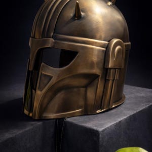 K&ouml;nnte beinhalten: Ein bronzefarbener Helm mit einem markanten Design, der ein Visier und zwei spitze Vorspr&uuml;nge auf der Oberseite aufweist. Der Helm hat einen metallischen Glanz und wird auf einer dunklen, strukturierten Oberfl&auml;che pr&auml;sentiert, was auf ein Sammler- oder Dekorationsst&uuml;ck hindeutet.
