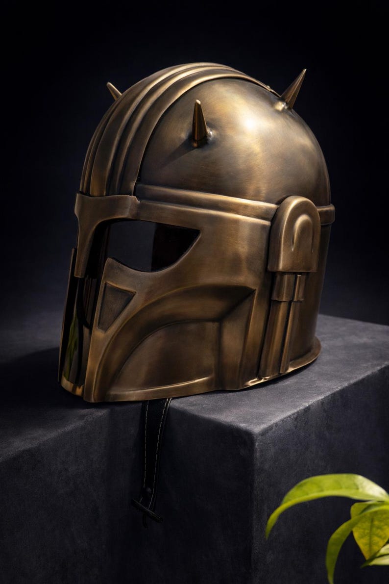 K&ouml;nnte beinhalten: Ein bronzefarbener Helm mit einem markanten Design, der ein Visier und zwei spitze Vorspr&uuml;nge auf der Oberseite aufweist. Der Helm hat einen metallischen Glanz und wird auf einer dunklen, strukturierten Oberfl&auml;che pr&auml;sentiert, was auf ein Sammler- oder Dekorationsst&uuml;ck hindeutet.