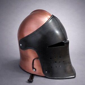 May include: A copper-colored medieval helmet with a black visor and face guard. The helmet has a rounded top and a riveted construction. The visor features small eye slits and ventilation holes. A leather strap with a buckle is attached to the helmet.